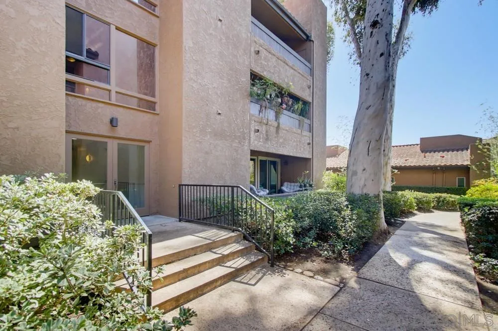 Condominium exterior with private entry steps, patio railing, shrubs, and a shaded walkway.