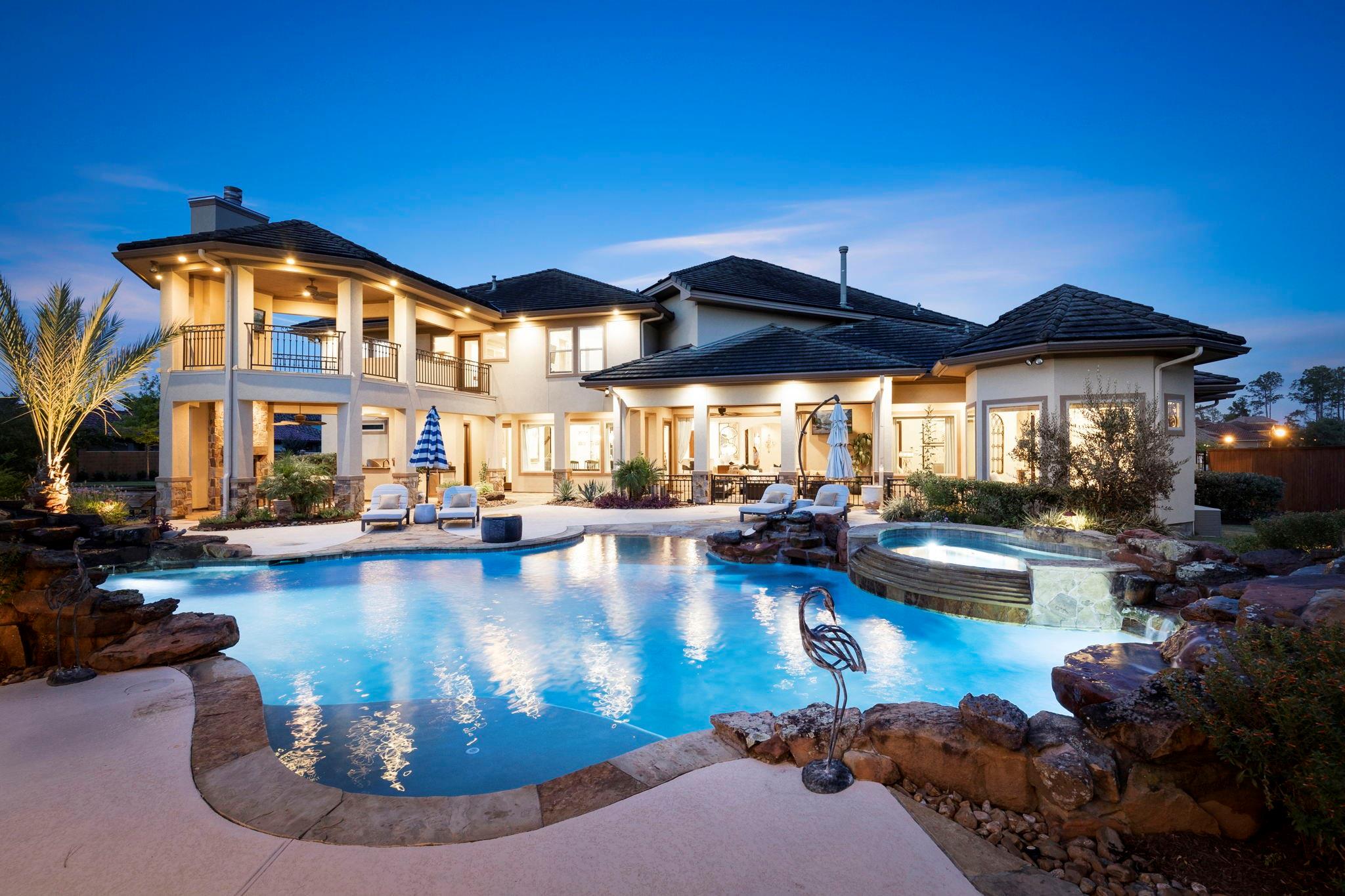 Large home behind a curving pool and raised spa, with illuminated patios at dusk.