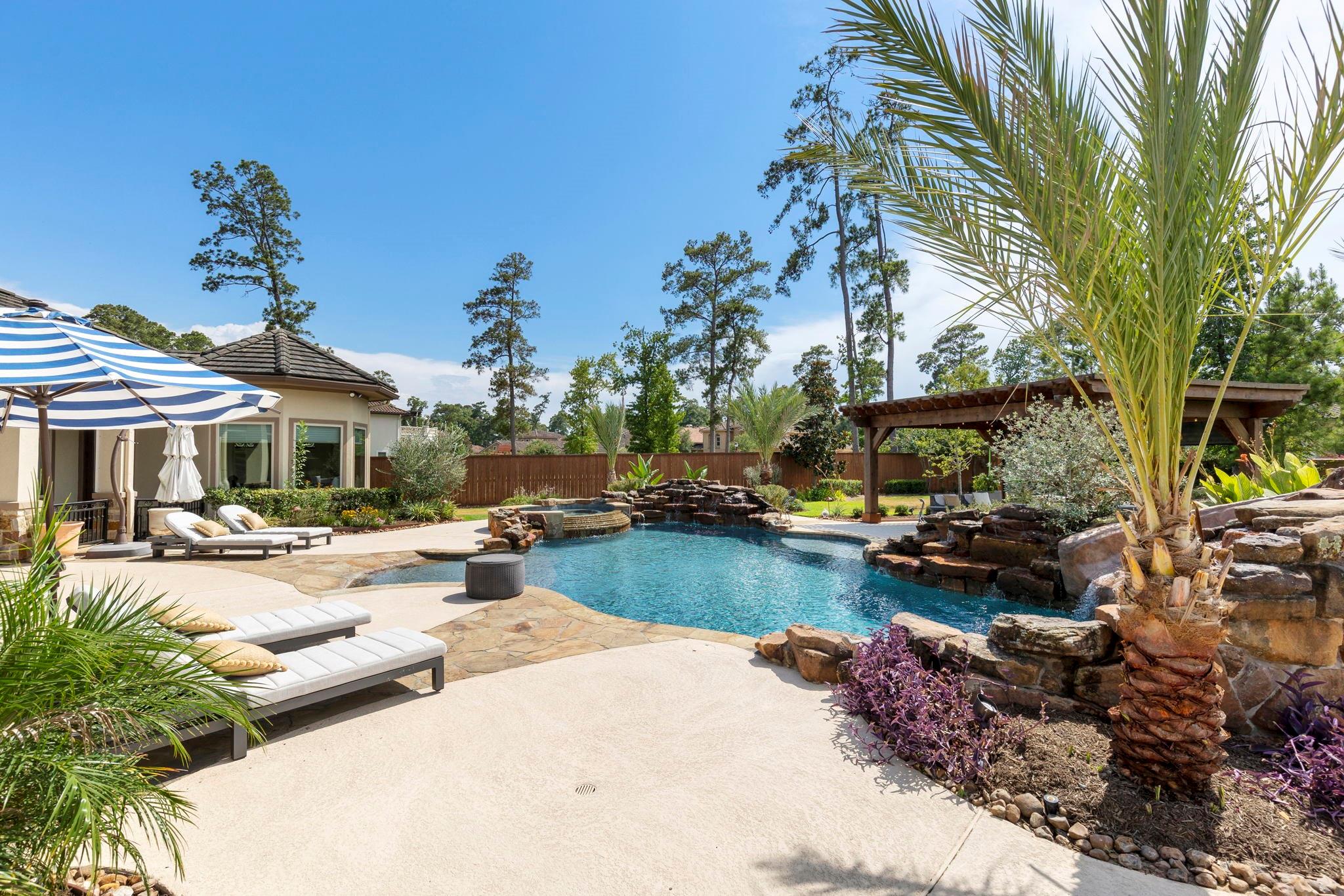 Backyard pool with a stone waterfall, landscaped rock borders, loungers, striped umbrellas, and a wooden pergola.