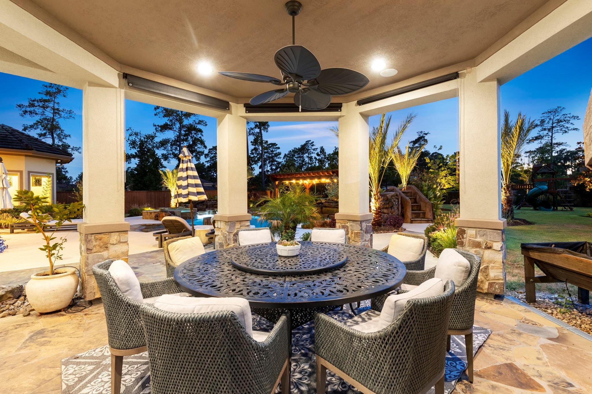 Covered patio with round dining table, wicker chairs, and a landscaped pool area beyond.