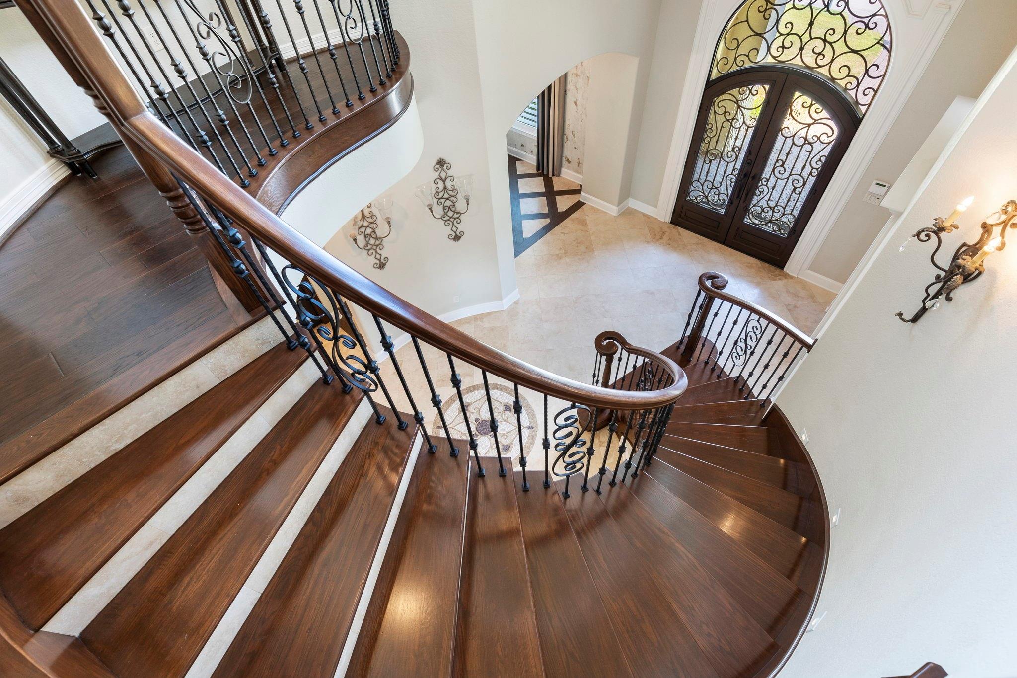 Grand foyer with a sweeping dark wood staircase and ornate iron railings beside glass-paneled double doors.