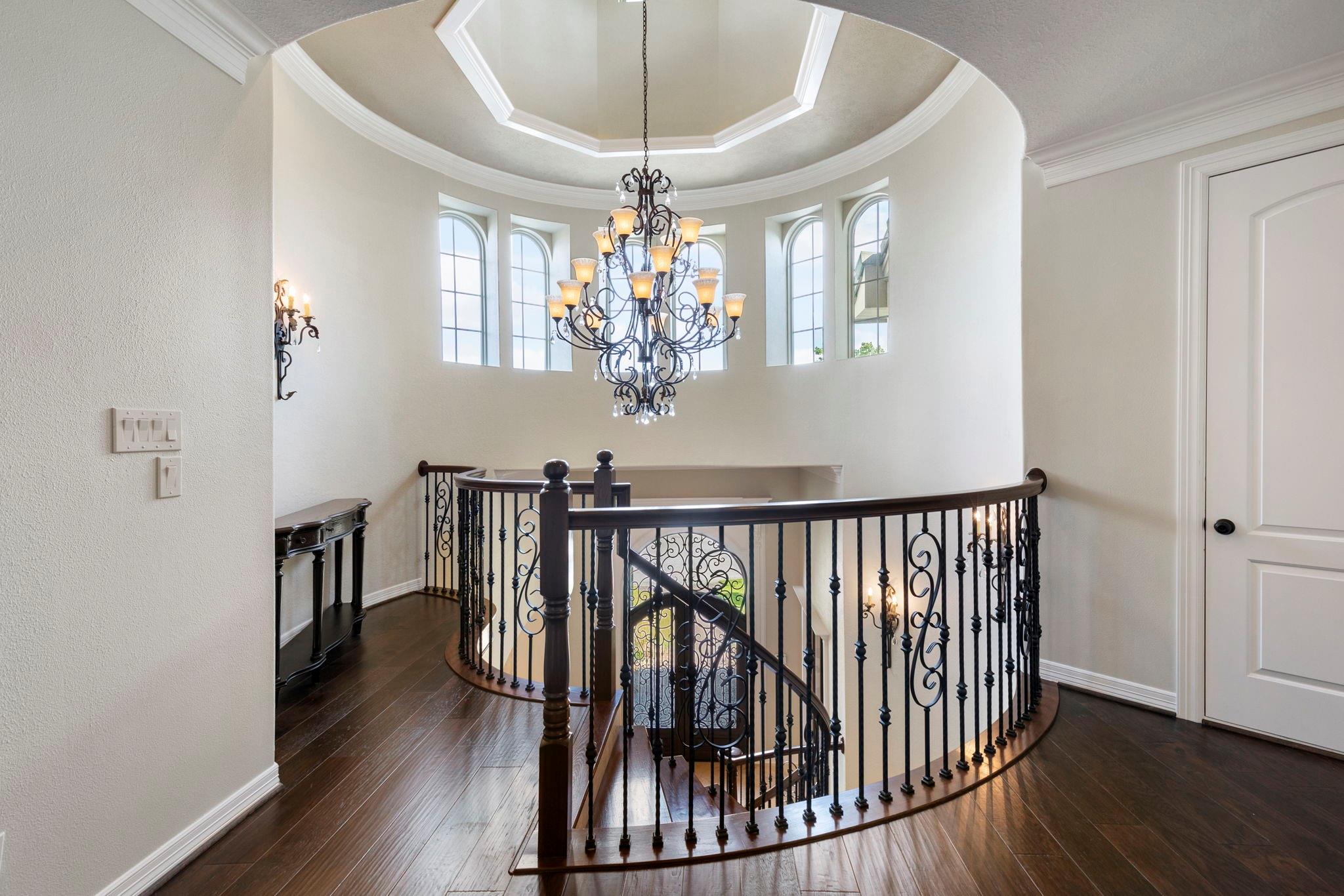 Upstairs landing with curved wrought-iron staircase railing, tall arched windows, and an ornate chandelier.