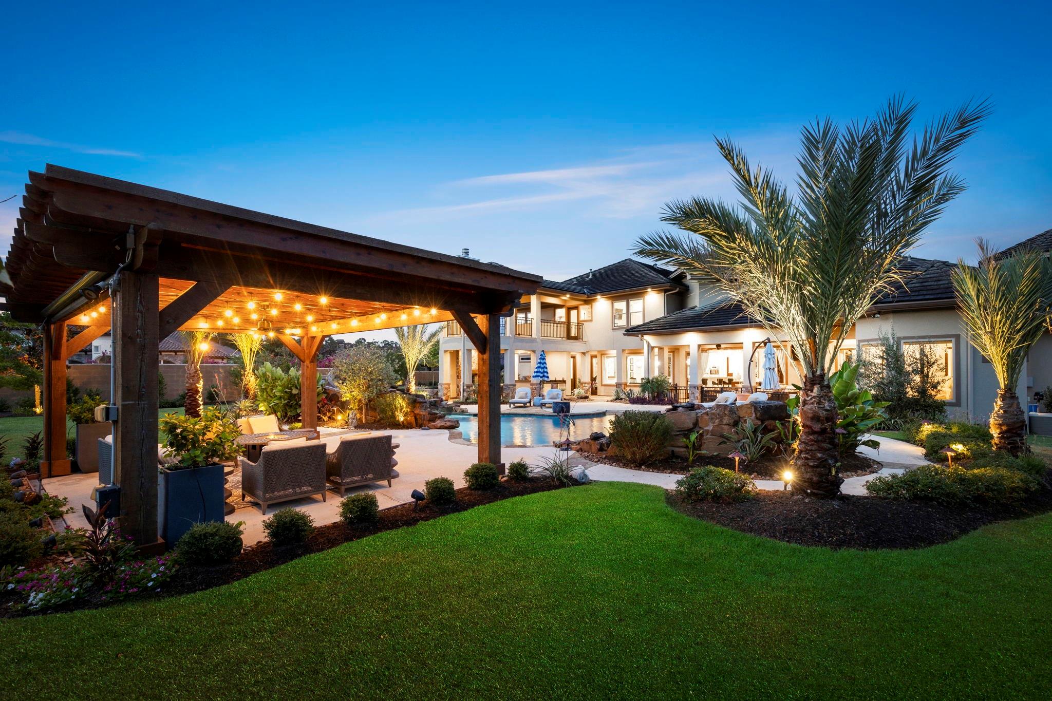 Backyard with a swimming pool, warmly lit pergola, palm trees, and landscaped lawn at dusk.