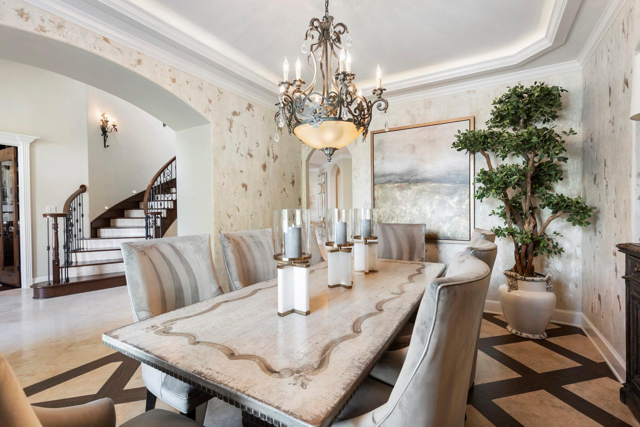 Dining room with a long stone table, ornate chandelier, and curved staircase.