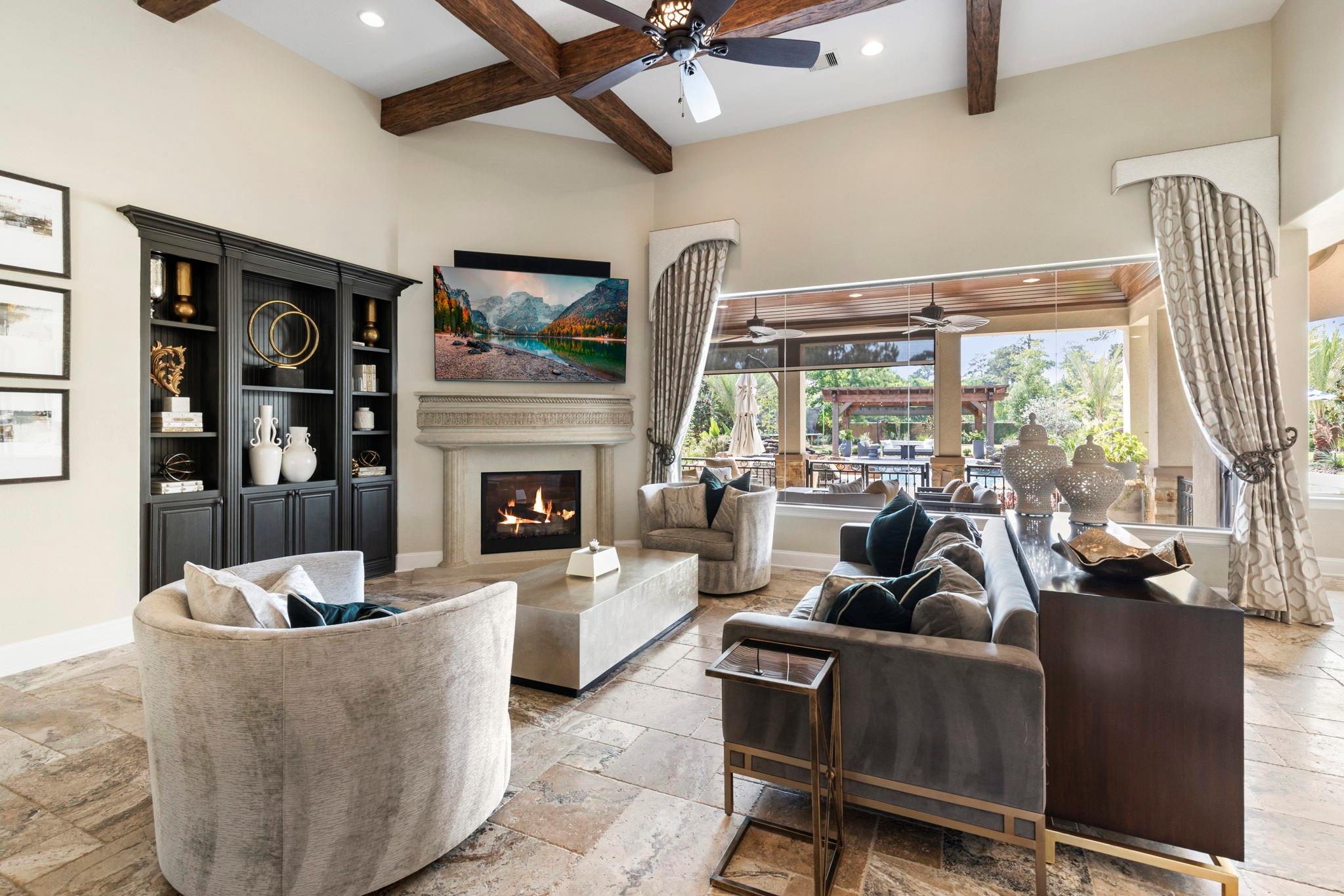 Staged living room with exposed wood beams, a stone fireplace, and windows overlooking a covered patio.