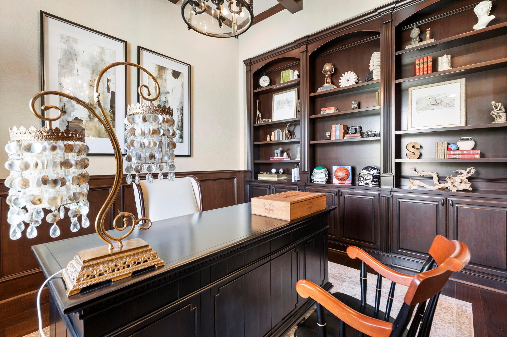 Home office with dark built-in bookshelves, a large desk, crystal lamps, and sports memorabilia.