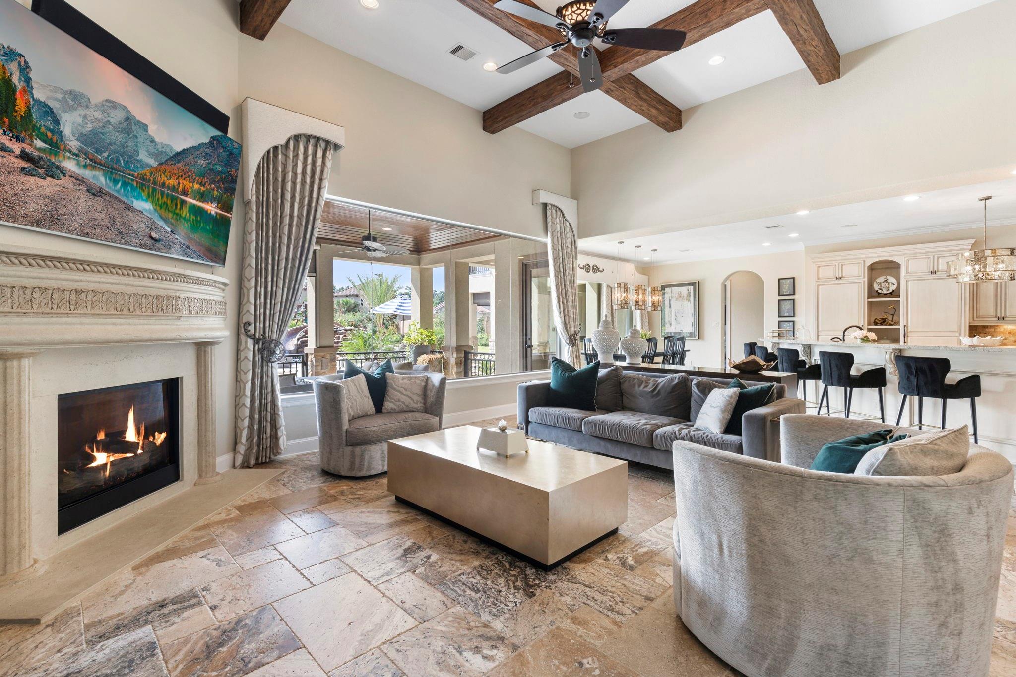 Living room with a stone fireplace, vaulted beamed ceiling, and large windows opening to a patio.