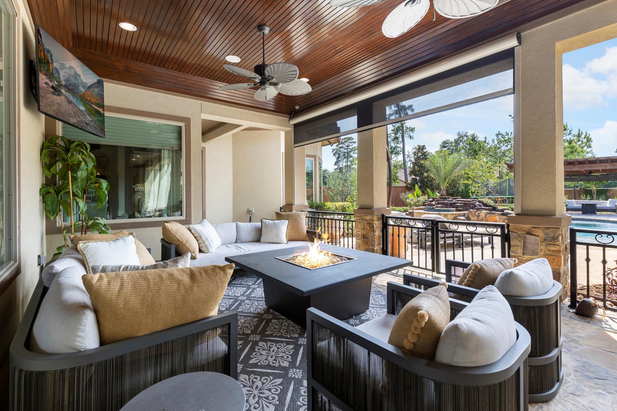 Covered patio with a wood ceiling, cushioned seating, fire table, and pool views.