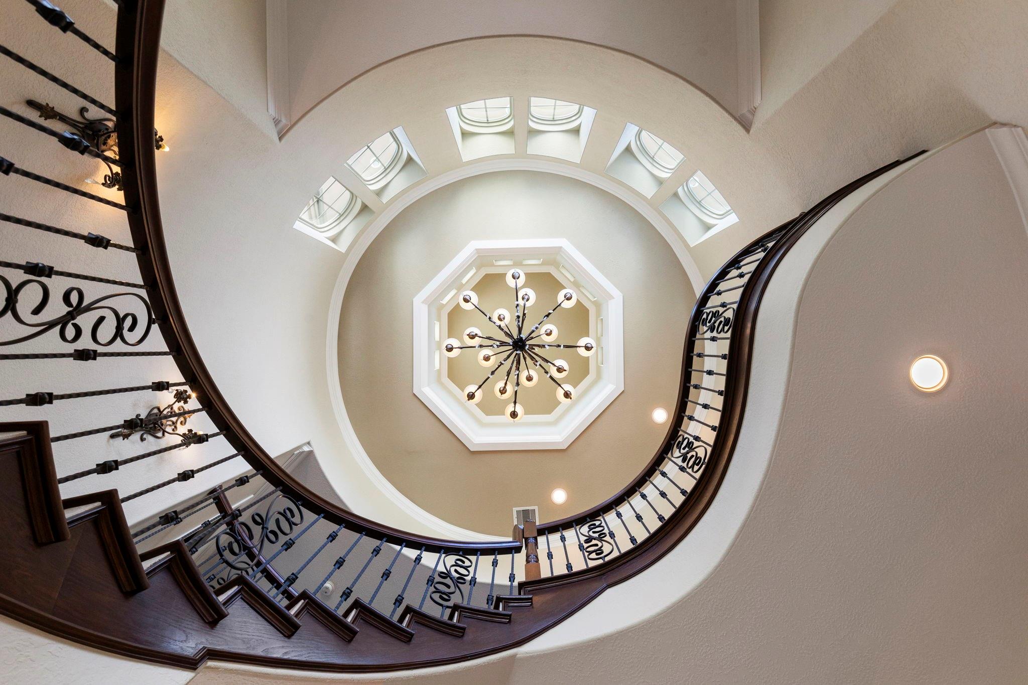 Circular stairwell with curved wood stairs, wrought-iron railings, skylights, and a central chandelier.