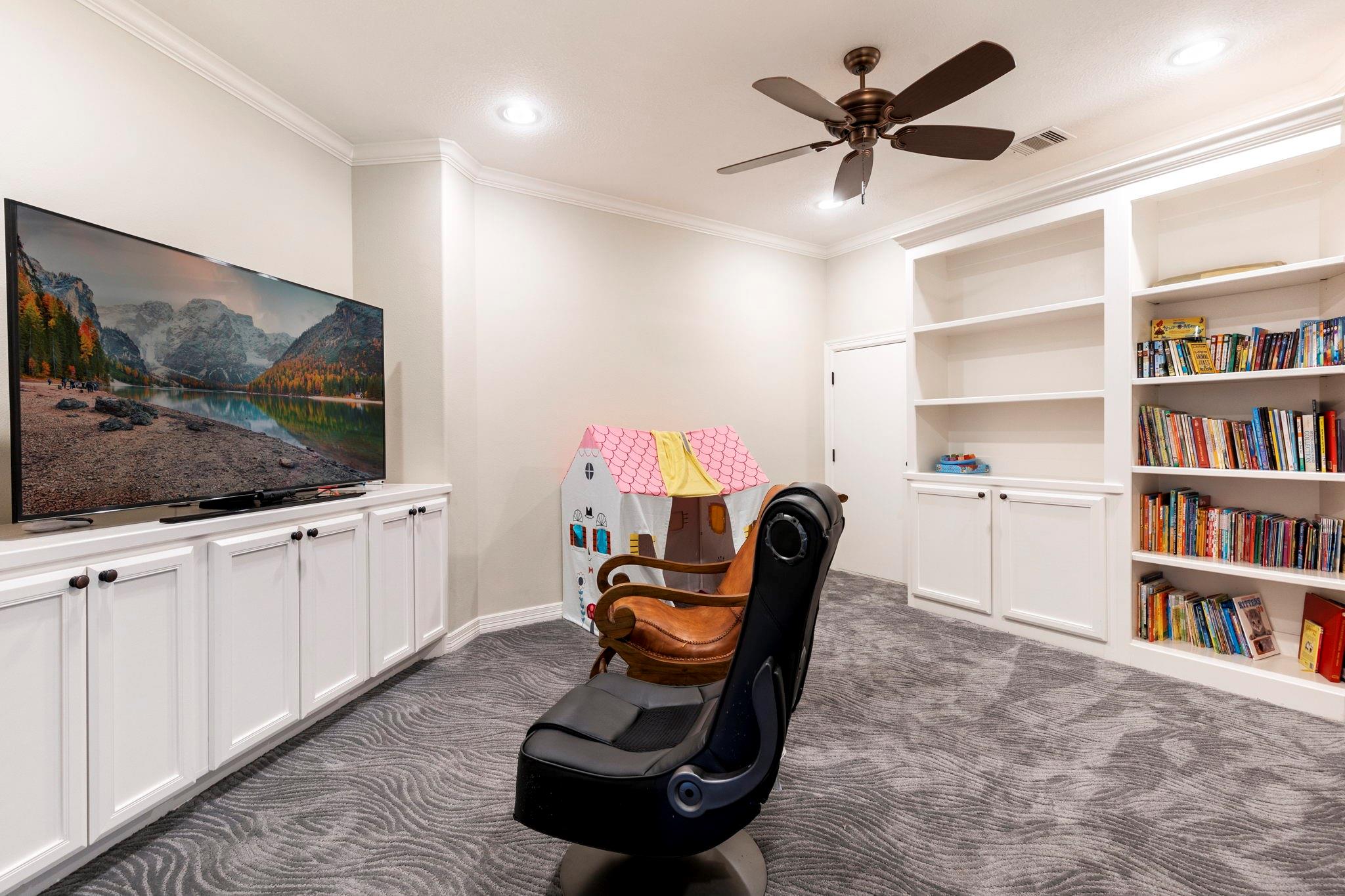 Media room with built-in bookshelves, a large television, massage chair, and children’s playhouse.