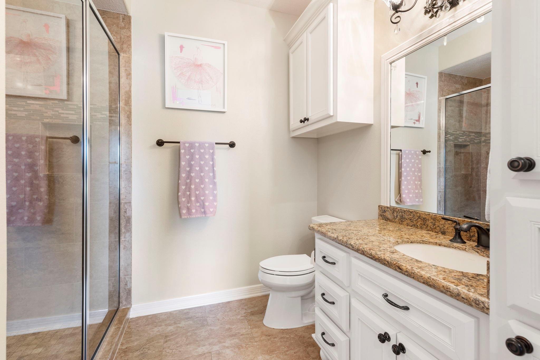 Bathroom with glass-enclosed tiled shower, granite vanity, white cabinetry, and a heart-patterned towel.
