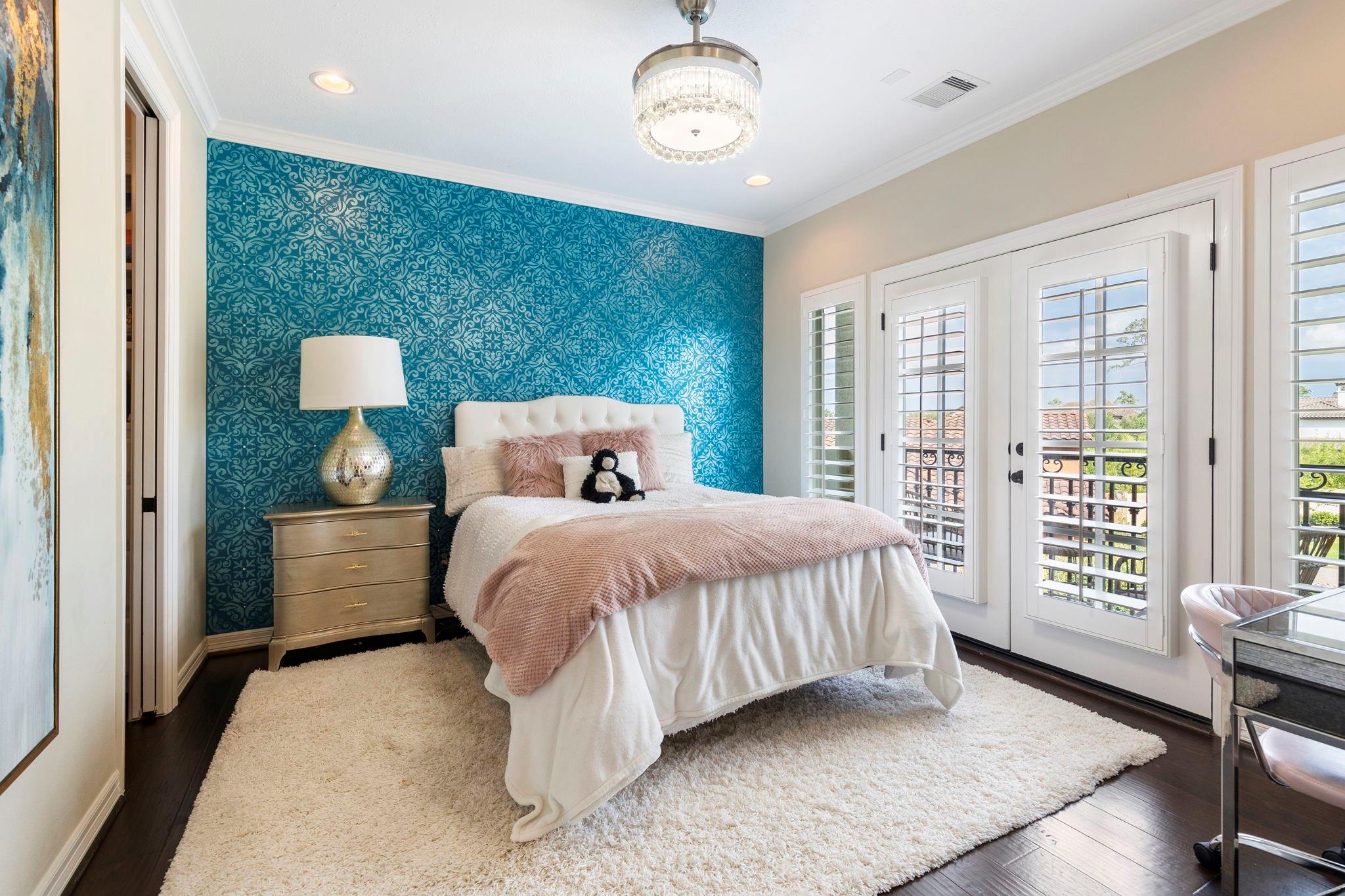 Bedroom with a teal patterned accent wall, blush bedding, and tall shuttered balcony doors.
