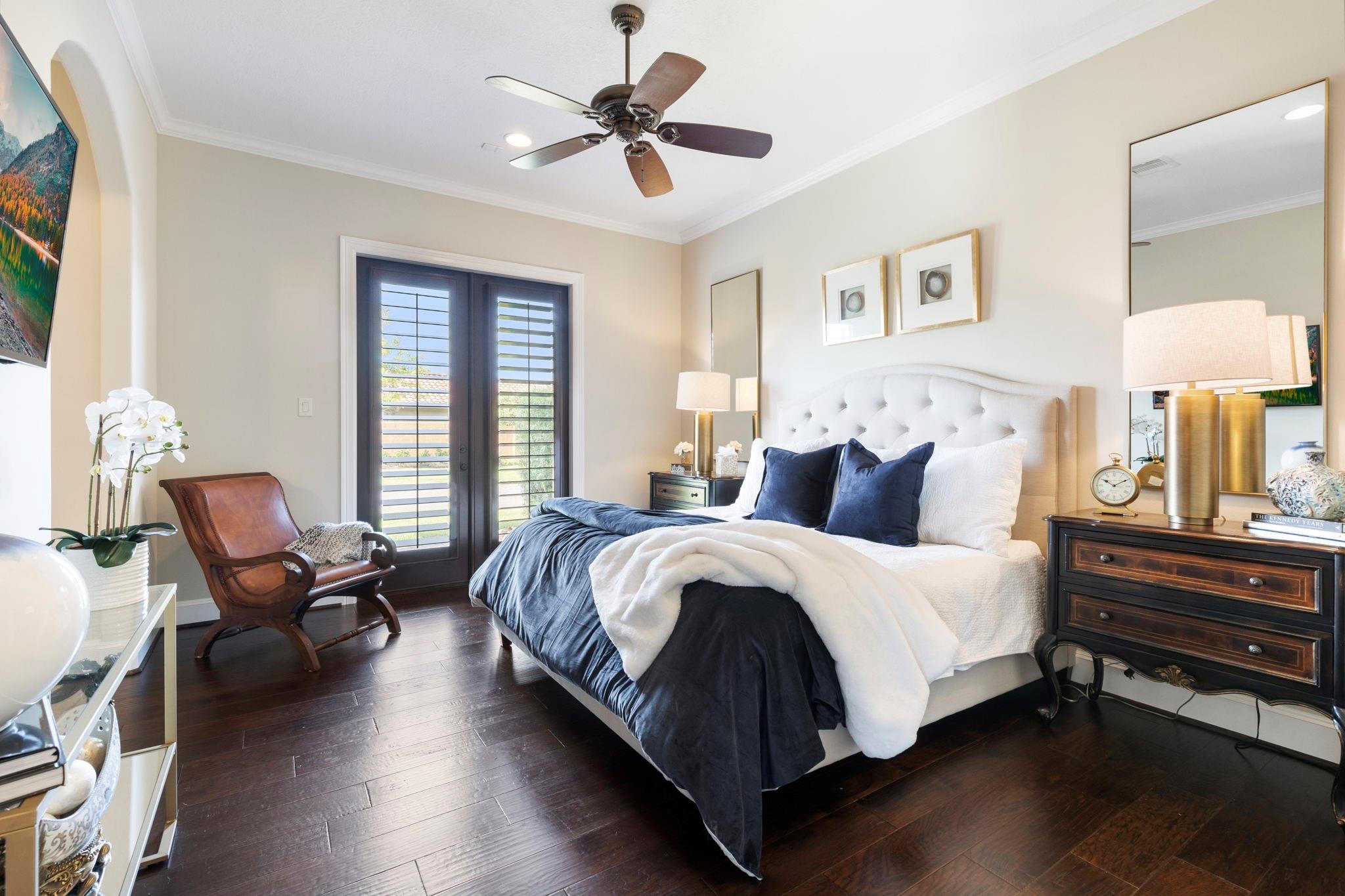 Bedroom with a tufted white bed, navy bedding, dark hardwood floors, and plantation-shuttered glass doors.