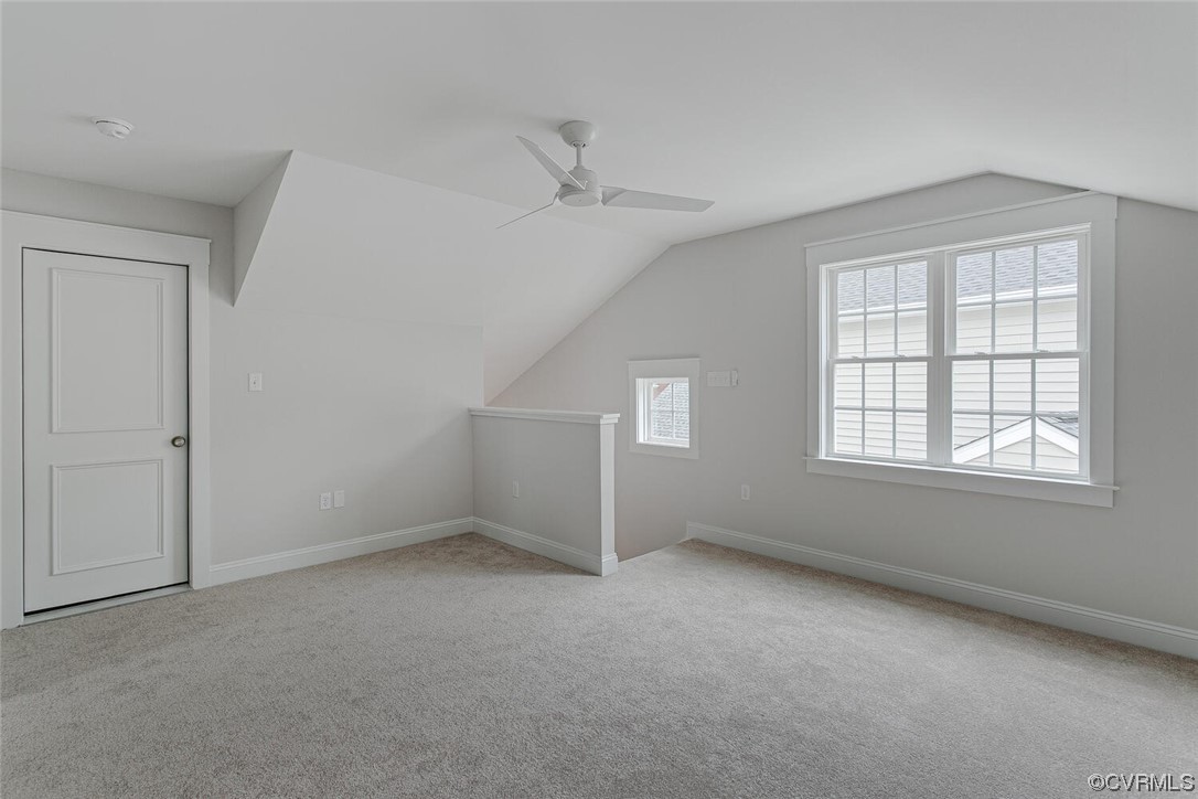Empty upstairs room with sloped ceilings, beige carpet, ceiling fan, and two windows.