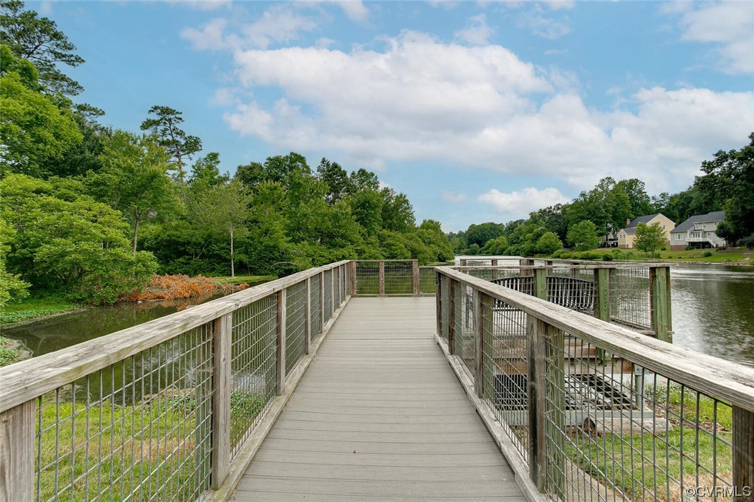 Wooden walkway extends over a lake, with tree-lined shore and houses across the water.
