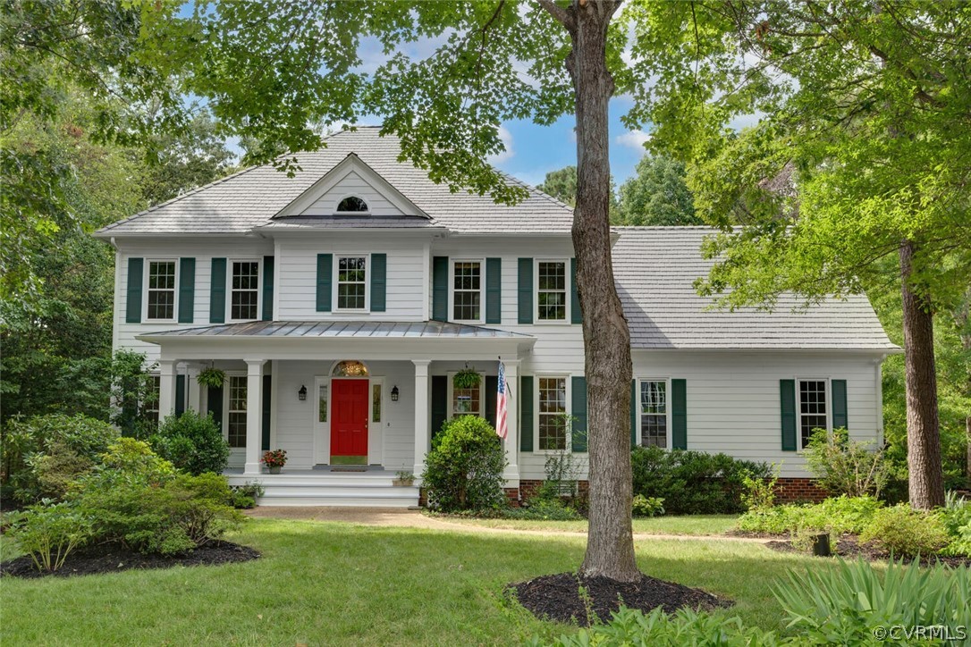 White two-story home with green shutters, a red front door, and a covered porch framed by mature trees.