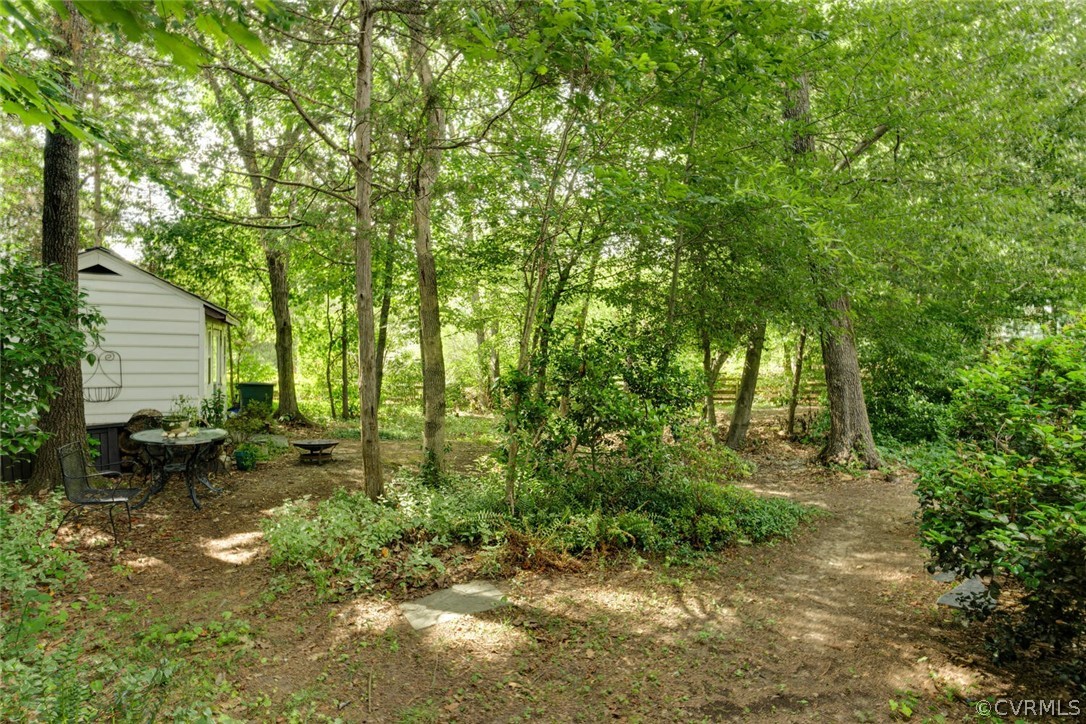 Wooded backyard with a dirt path and a small patio seating area beside a white-sided structure.