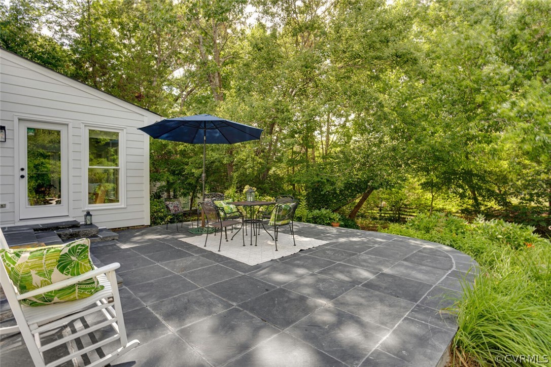 Slate patio with umbrella-shaded dining set beside a white-sided home and leafy garden.