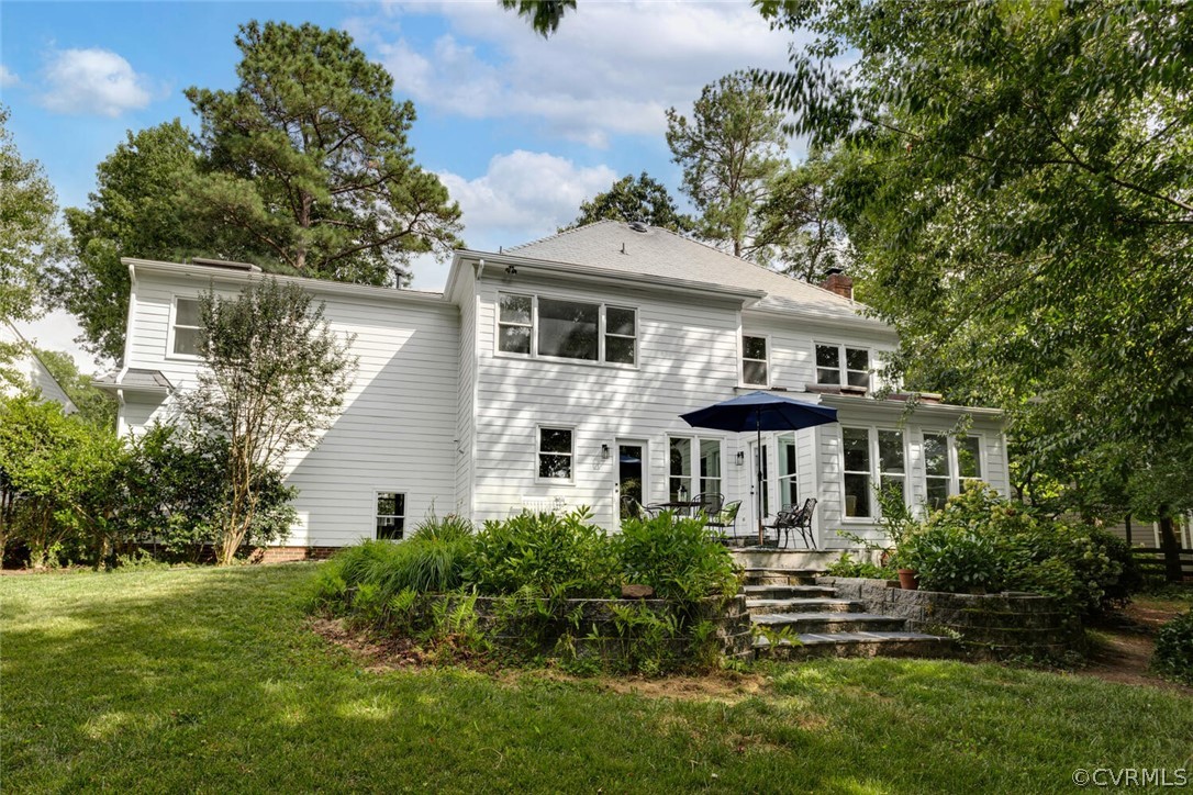 White two-story home with a raised stone patio, outdoor seating, and mature trees.