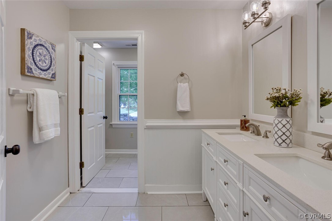 Bathroom with long white double vanity, tiled floor, and doorway to a windowed adjoining room.