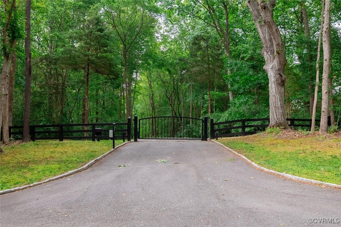 Paved driveway ending at a black metal gate, surrounded by mature trees and fencing.