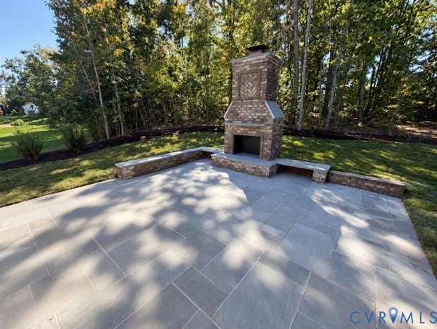 Stone patio with a brick fireplace and low seating walls beneath mature trees.