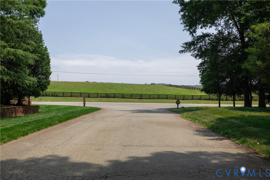 Paved driveway ending at a road beside a fenced grassy field, framed by mature trees.