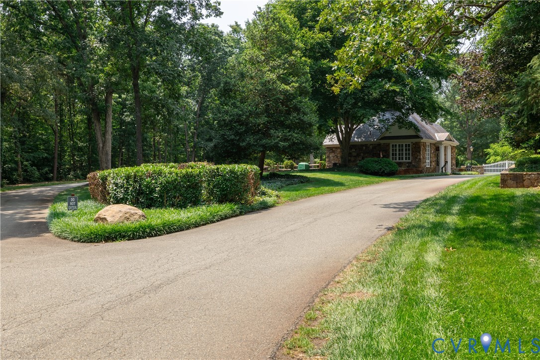Paved drive curves through mature trees toward a small stone building and white gate.