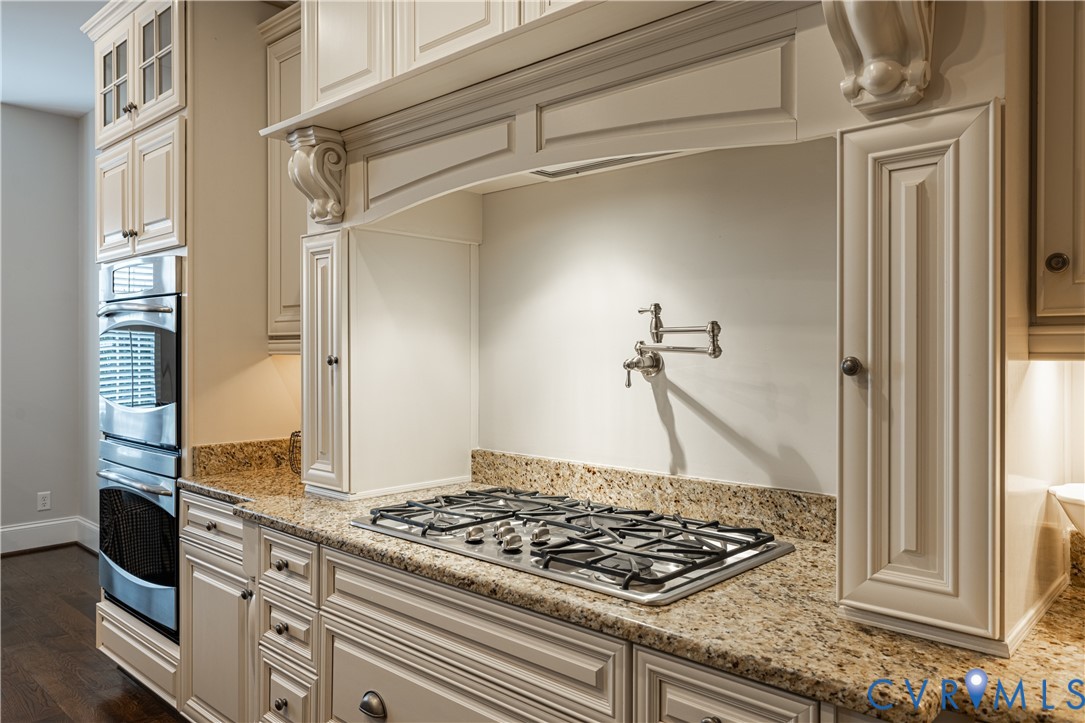 Kitchen cooktop with a pot filler, speckled stone counters, ornate cream cabinetry, and built-in double oven.