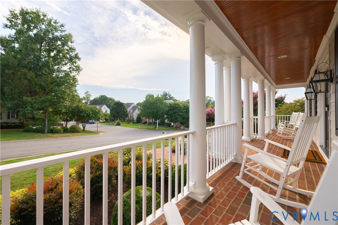 Covered front porch with white columns, brick flooring, and rocking chairs overlooking a tree-lined street.