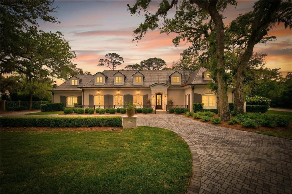 Large home at dusk with illuminated windows, mature trees, and a curved brick driveway.
