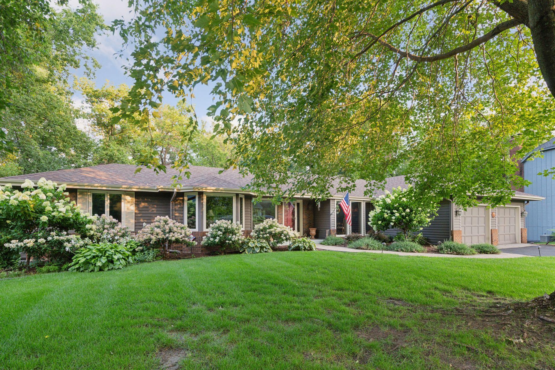 Single-story home with a two-car garage, flowering shrubs, and mature trees shading the front lawn.