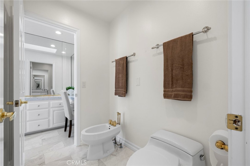 Bright bathroom with a toilet, bidet, brown towels, and an adjoining vanity visible through the doorway.
