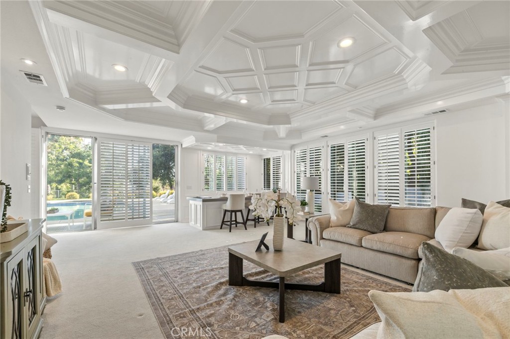 Living room with a coffered ceiling, plantation shutters, and sliding doors overlooking a pool.