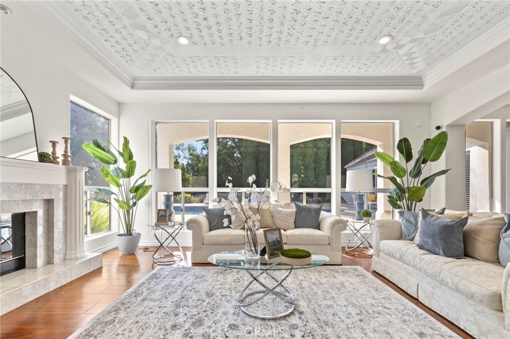 Staged living room with cream sofas, a marble fireplace, patterned ceiling, and windows overlooking a pool.