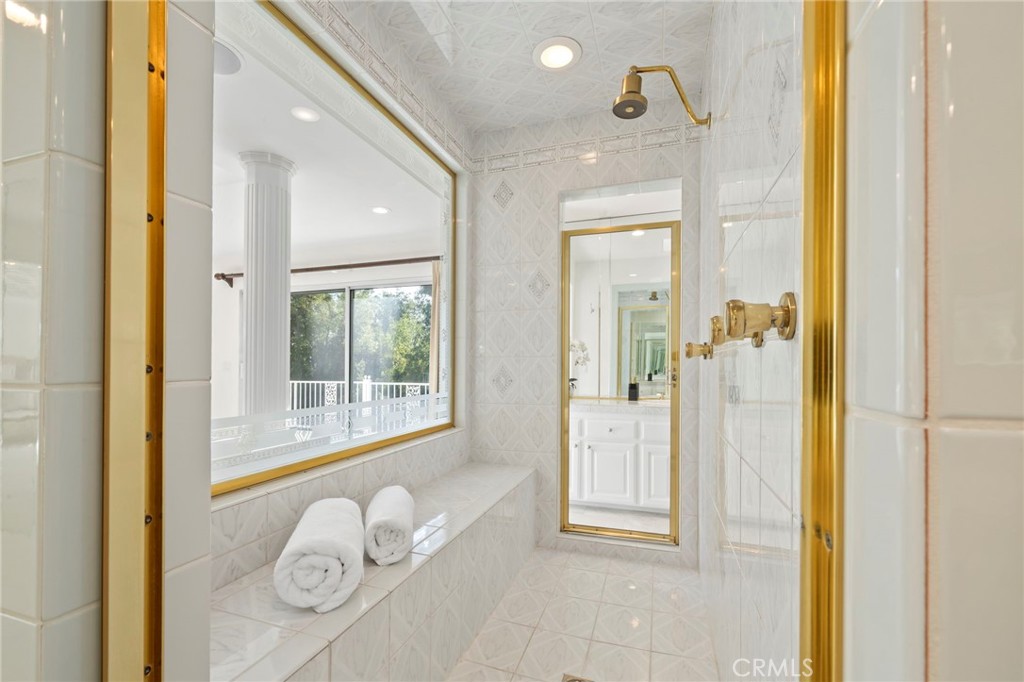 White-tiled shower with gold fixtures, a built-in bench, rolled towels, and a window overlooking greenery.