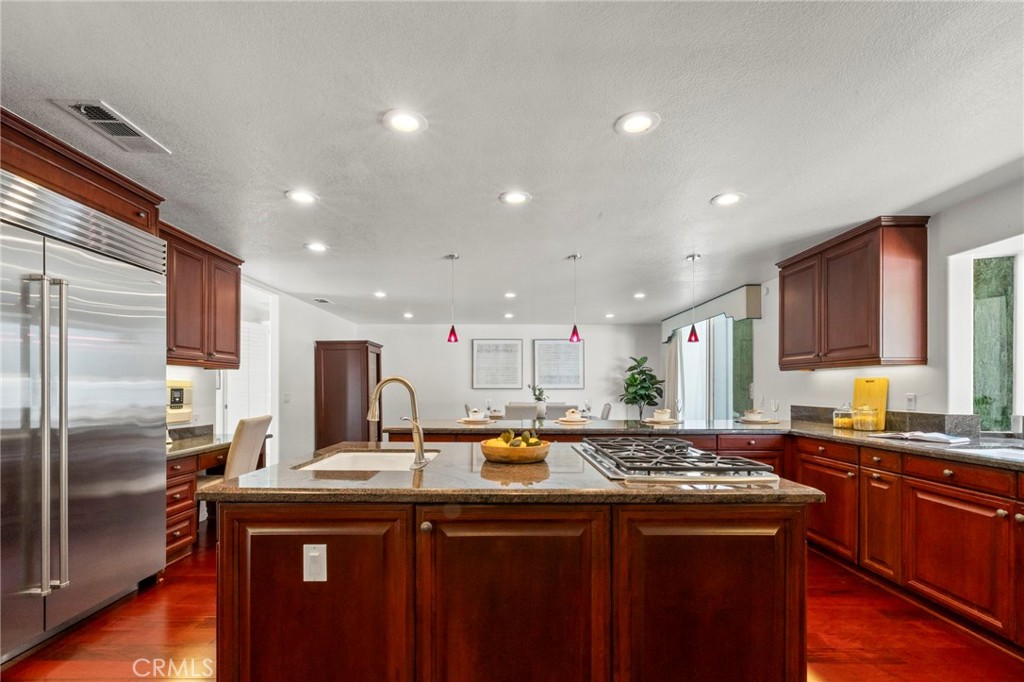 Kitchen with a granite island featuring a built-in cooktop and sink, dark wood cabinetry, and stainless steel appliances.