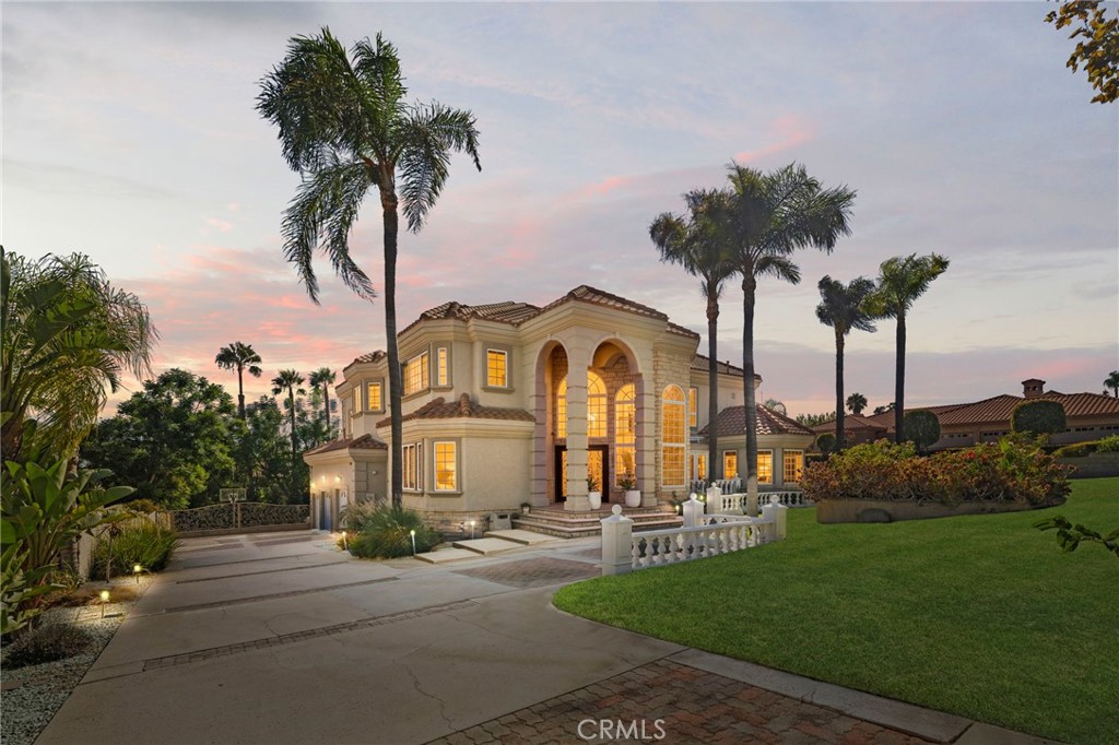 Mediterranean-style home with glowing arched windows, palm trees, and a broad driveway at dusk.