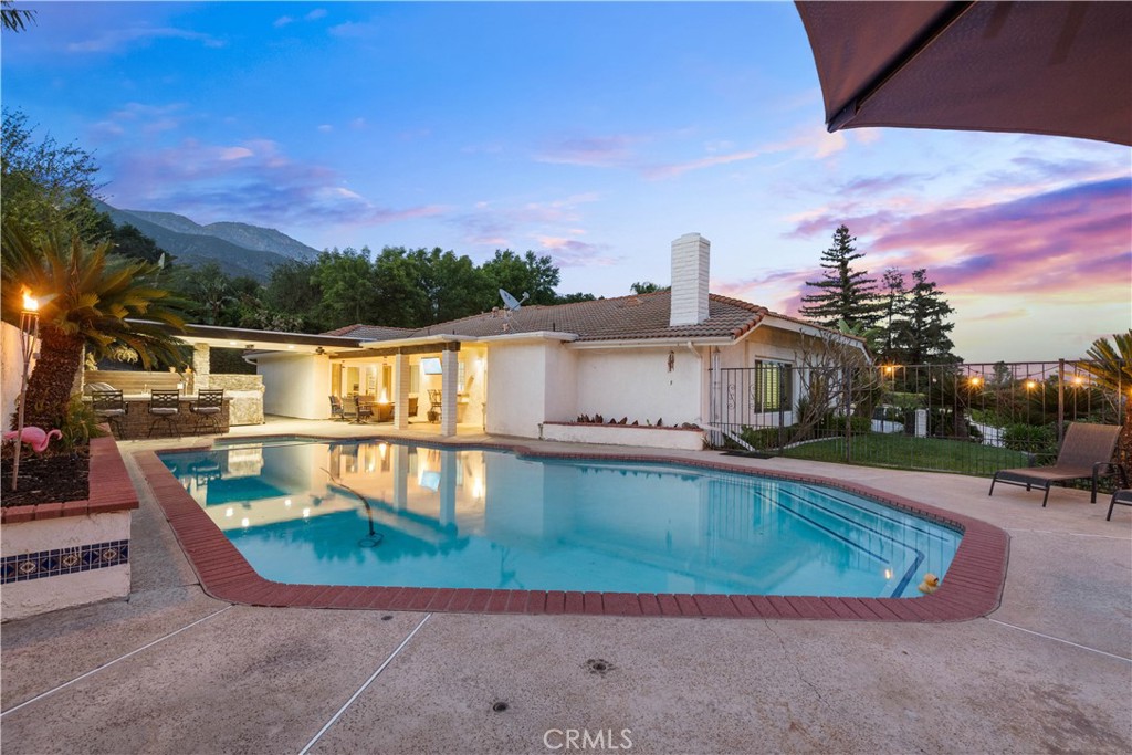 Backyard swimming pool beside a single-story home, with patio seating and mountains at dusk.