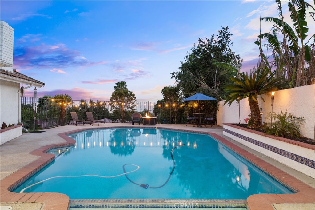 Backyard swimming pool with tiled edging, lounge chairs, and a fire pit at dusk.