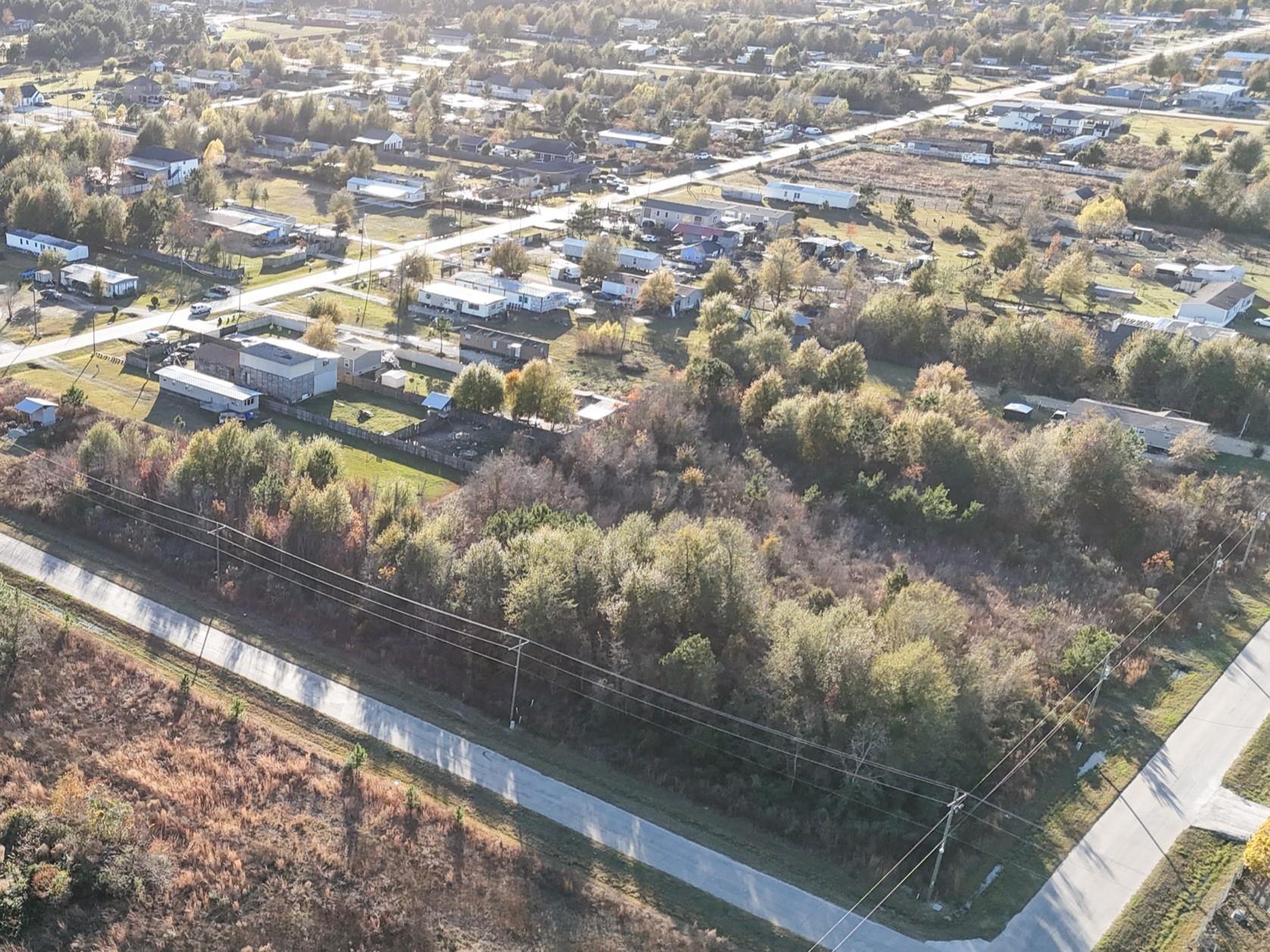 Aerial view of wooded vacant land beside paved roads and a neighborhood of scattered homes.