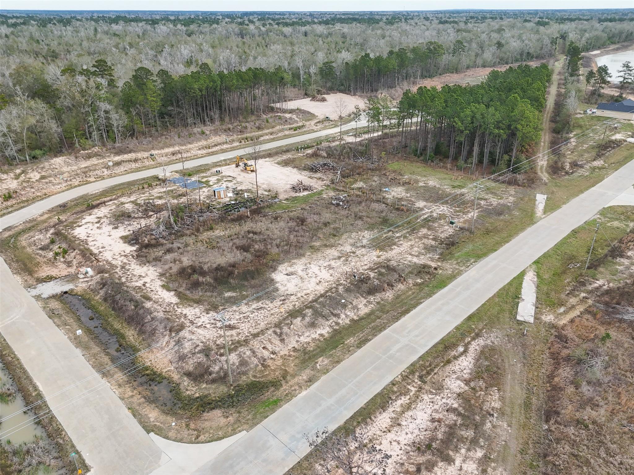 Aerial view of cleared sandy lots beside wooded land, with an excavator and paved roads.