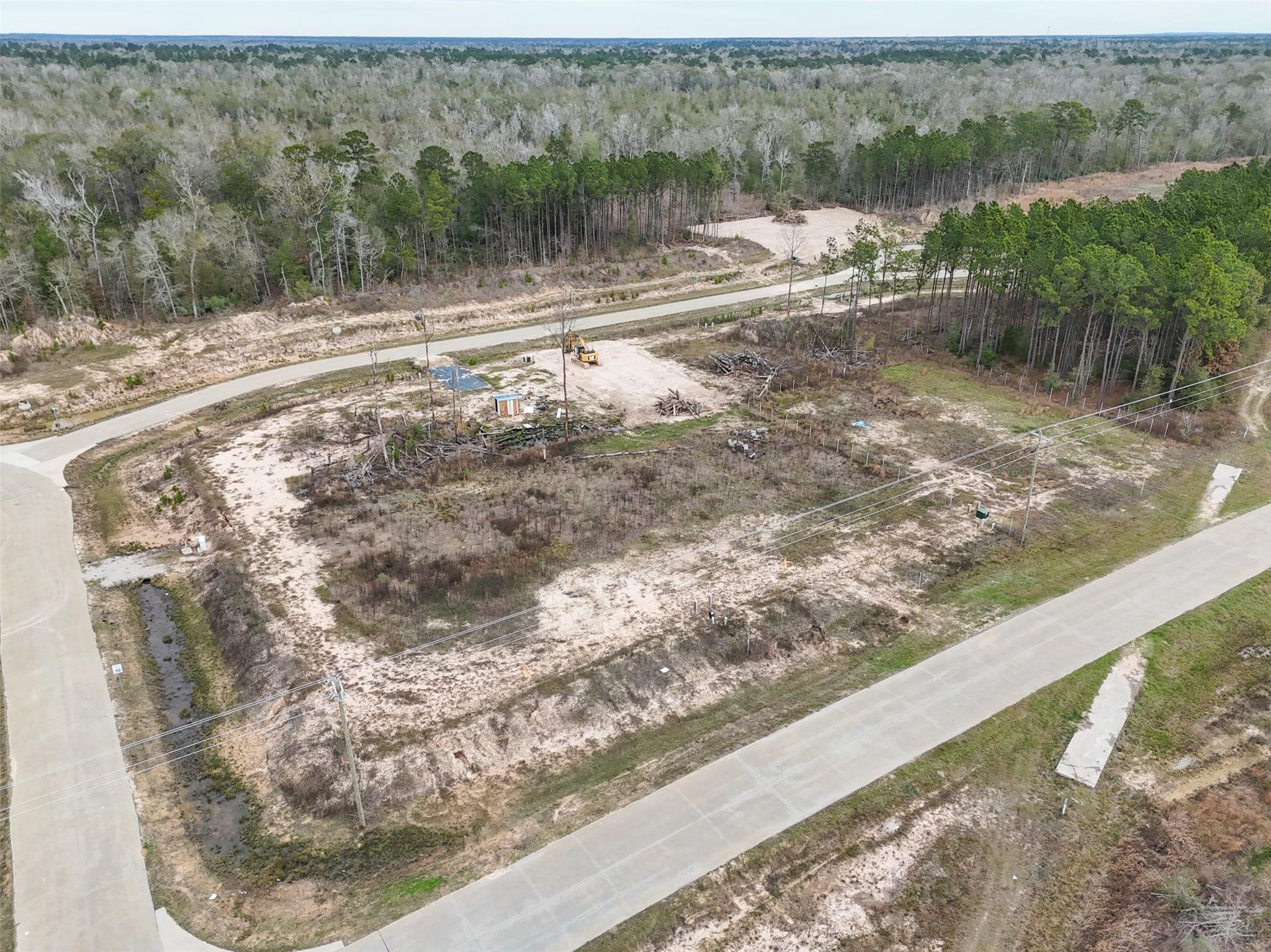 Aerial view of a sandy vacant lot bordered by roads and woods, with utility lines and an excavator.