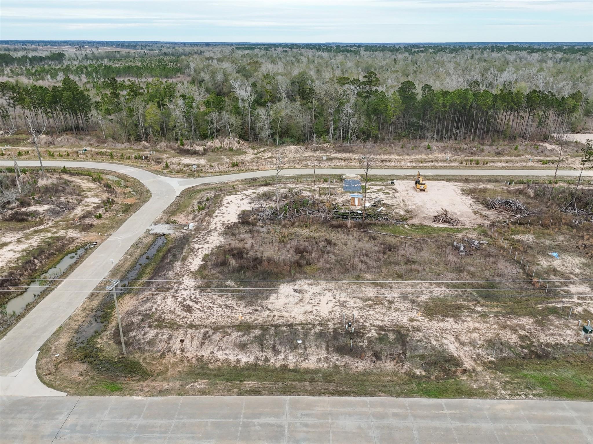 Aerial view of a cleared sandy lot beside a curving road, with an excavator and woodland beyond.