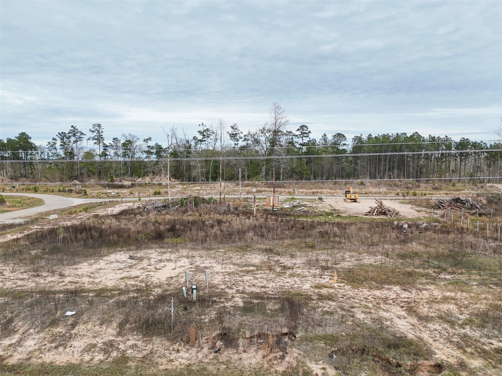 Aerial view of a cleared sandy lot with sparse brush, utility markers, and a curving road beside woods.