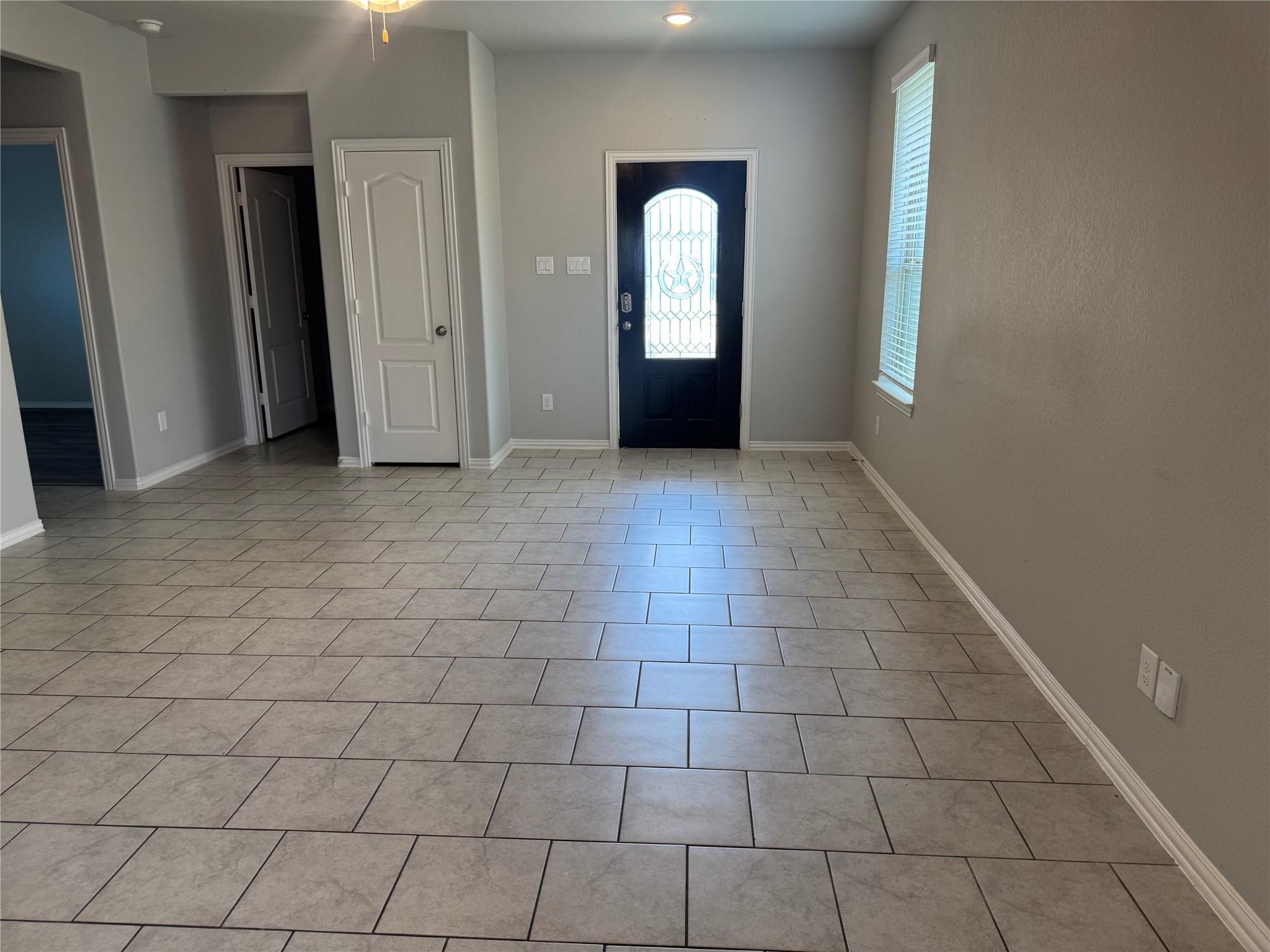 Empty entryway with large gray tile floors, a dark front door with decorative glass, and a window with blinds.