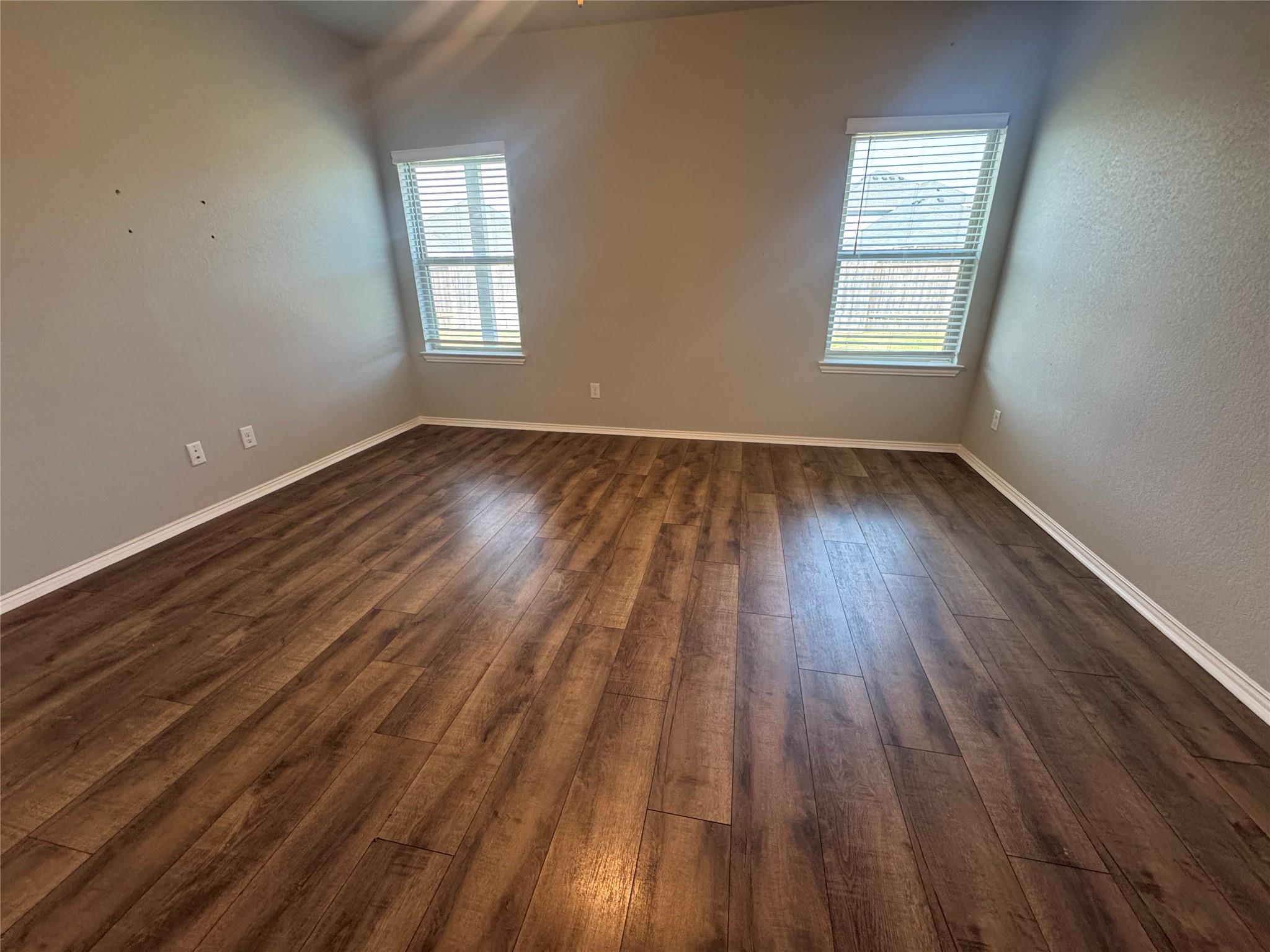 Empty room with brown plank flooring and two windows with blinds.