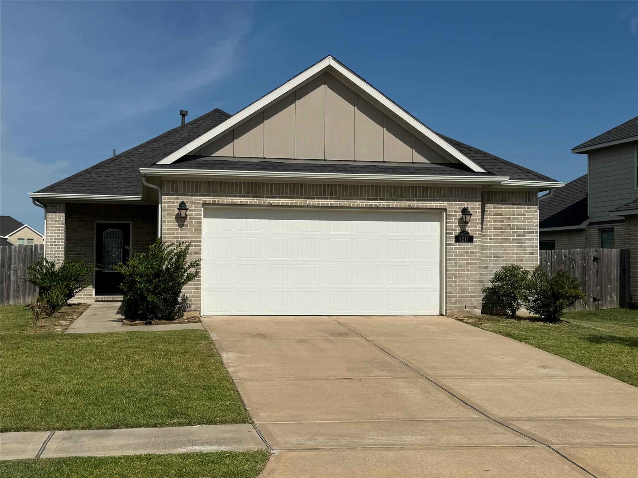 Single-story brick house with a white double garage door, front-facing gable, and broad concrete driveway.