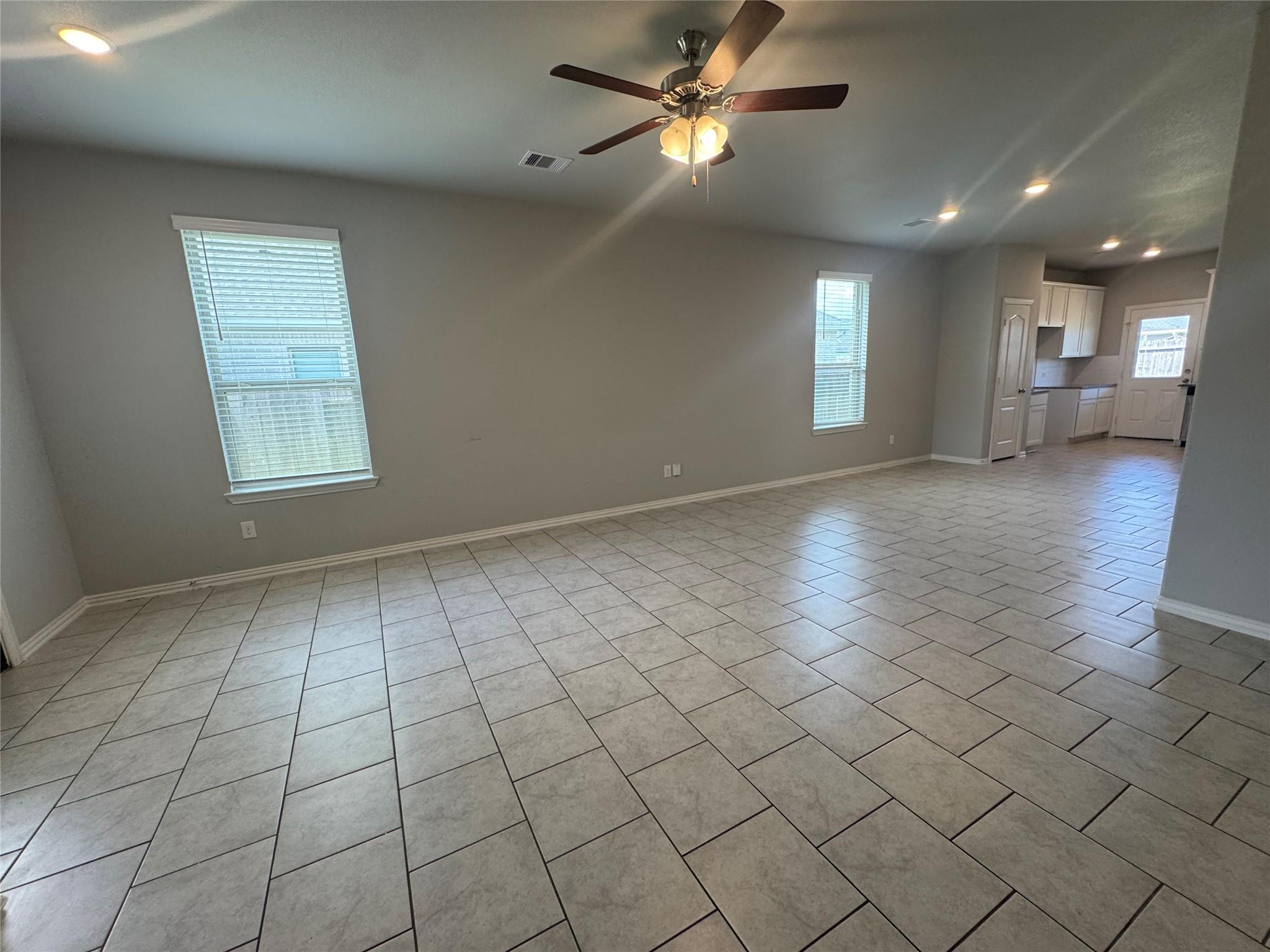 Unfurnished living area with large tile flooring, two blind-covered windows, and an open kitchen beyond.