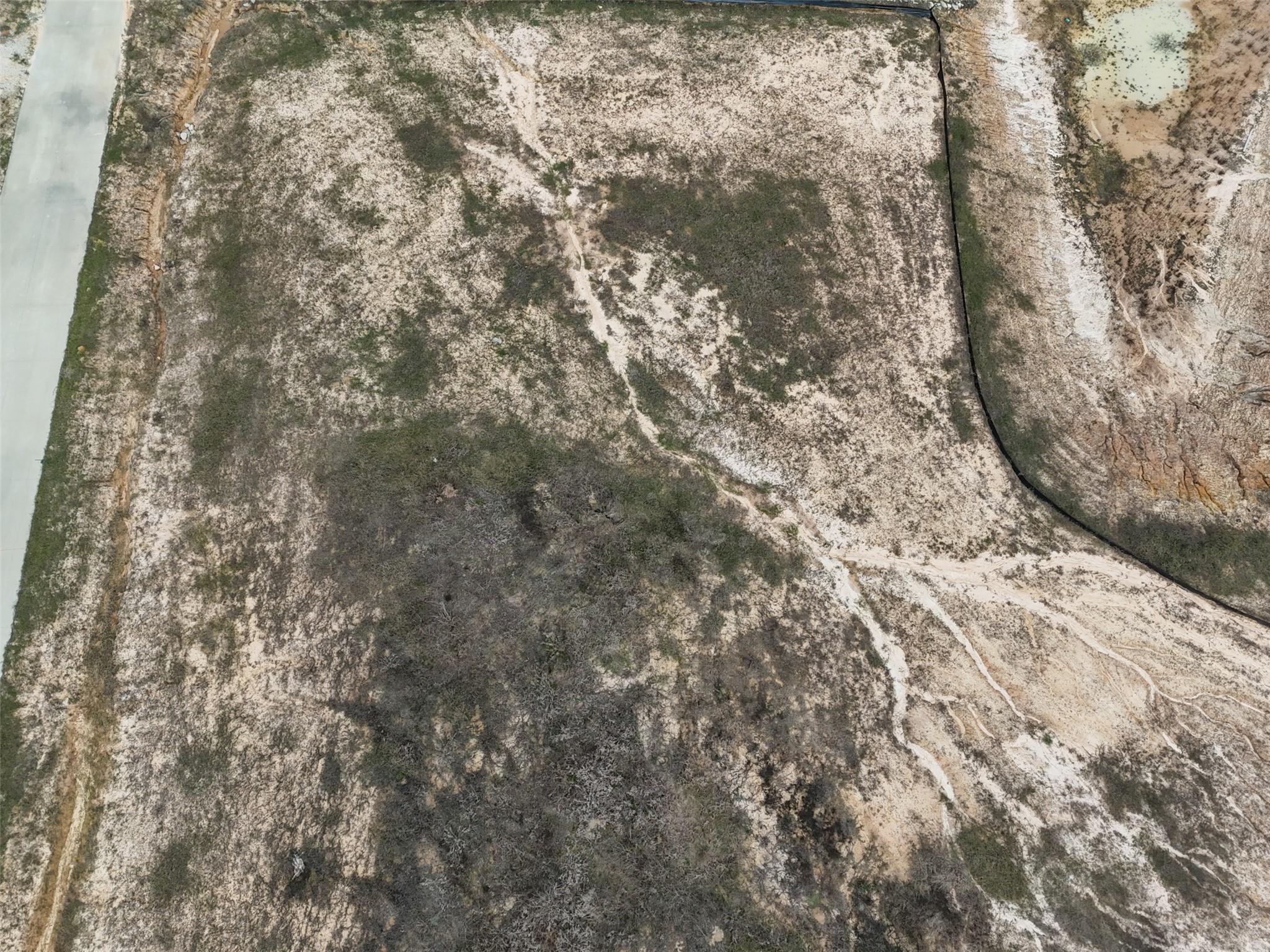 Aerial view of a sandy, brush-covered vacant lot with dirt tracks beside a paved road.